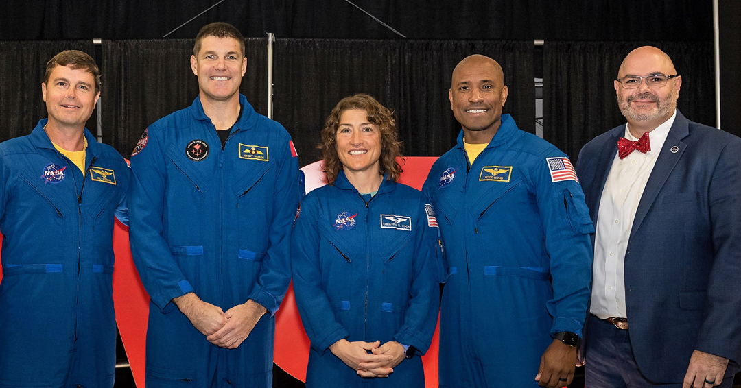 A man stands at a podium addressing attendees seated at round tables during an awards ceremony in a bright conference room.