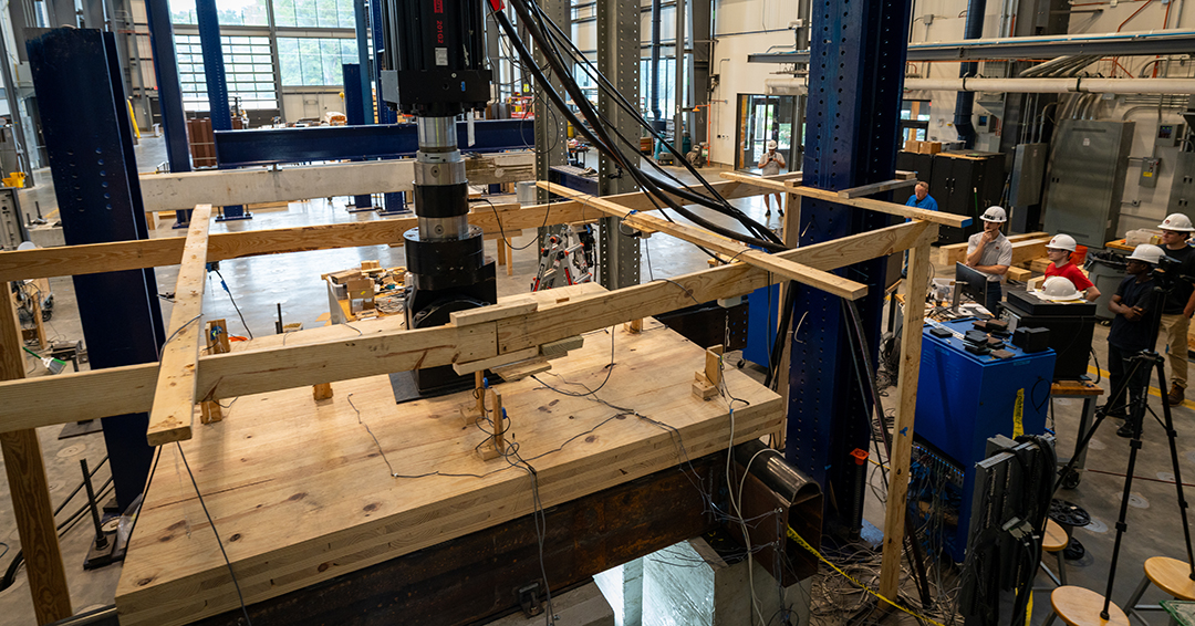 A large timber assembly is positioned beneath structural testing equipment in Auburn’s Advanced Structural Engineering Laboratory.