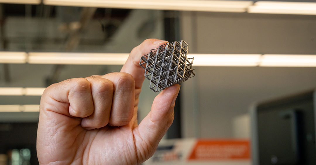 A hand holds a small, 3D-printed metal lattice structure.