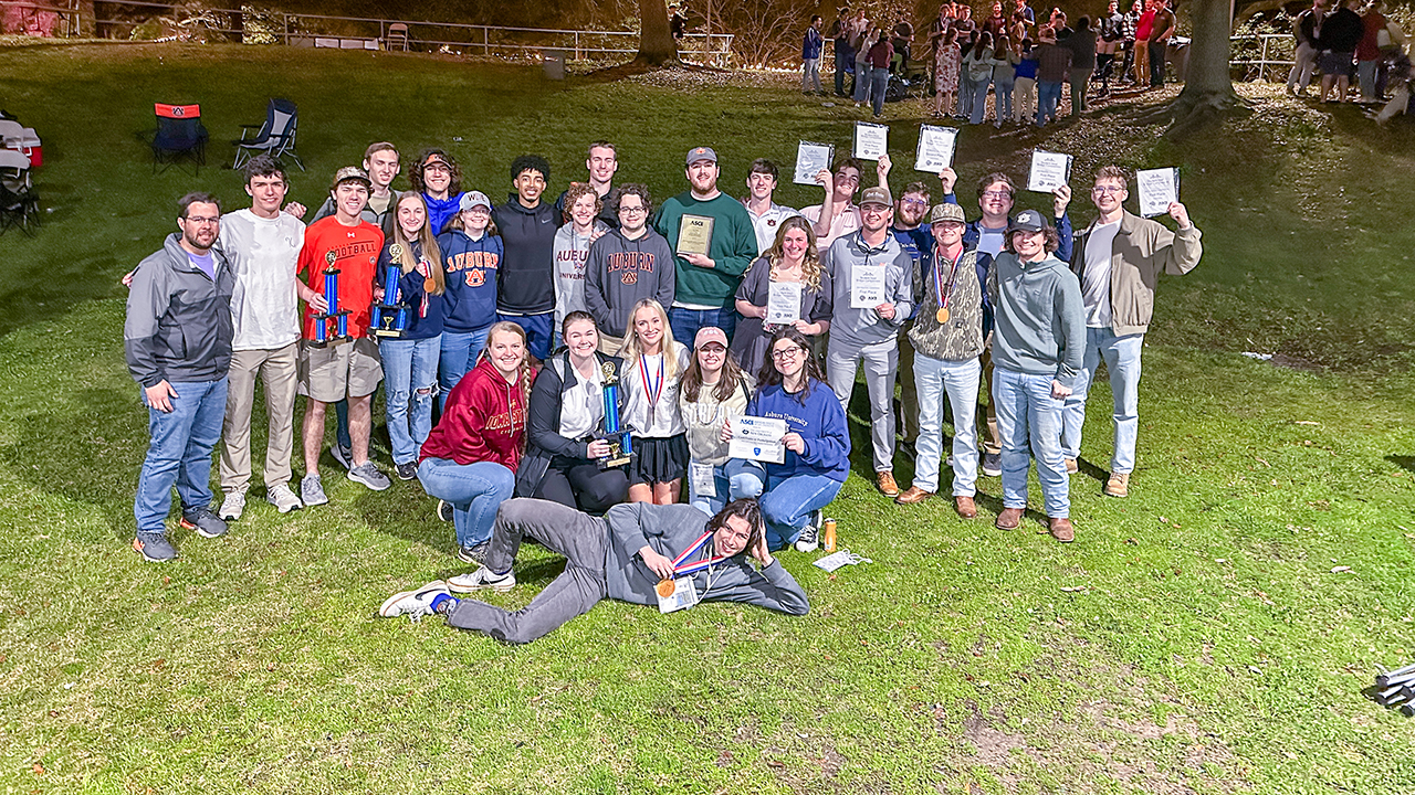  several students pose for a photo in a grassy field holding up awards and trophies 
