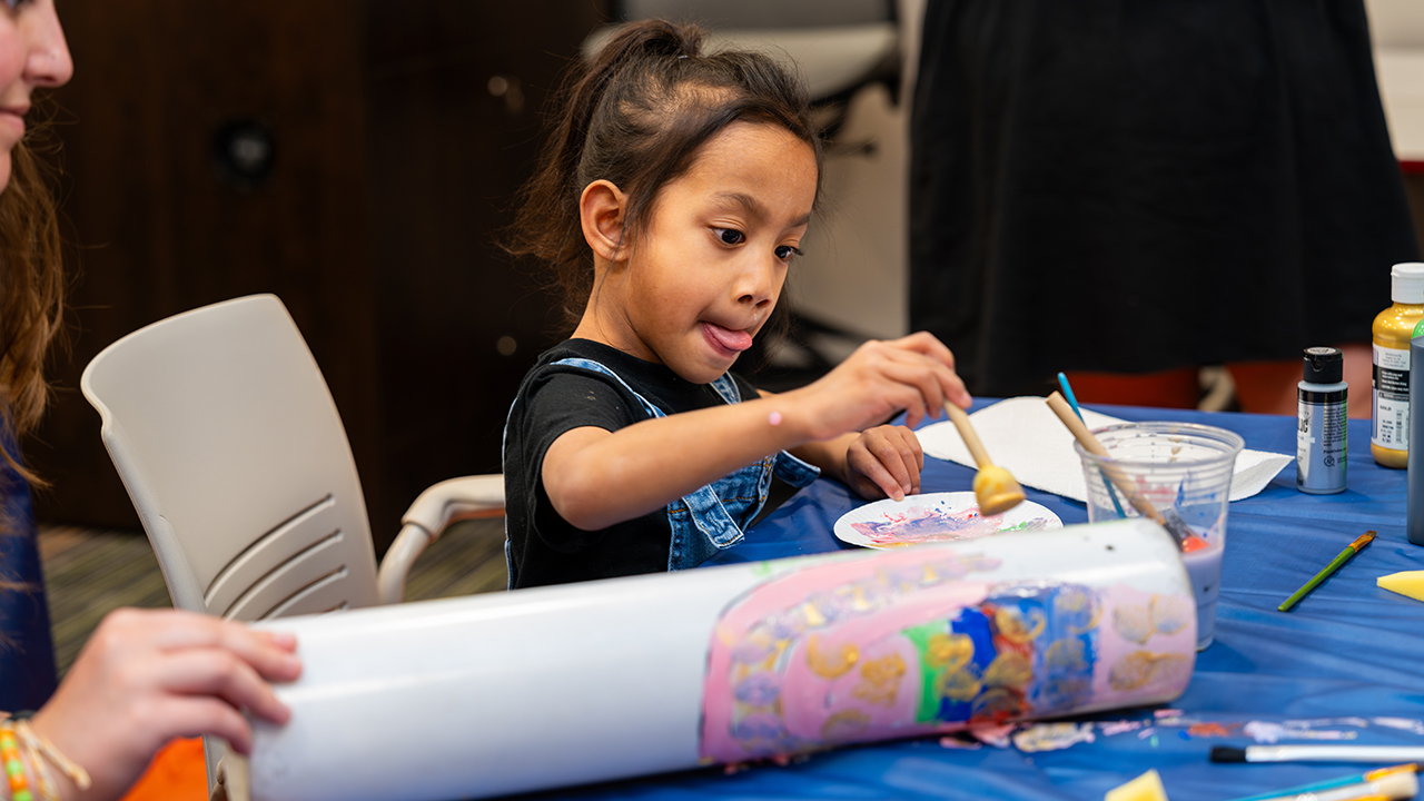 young girl in blue overalls paints a circular tube.