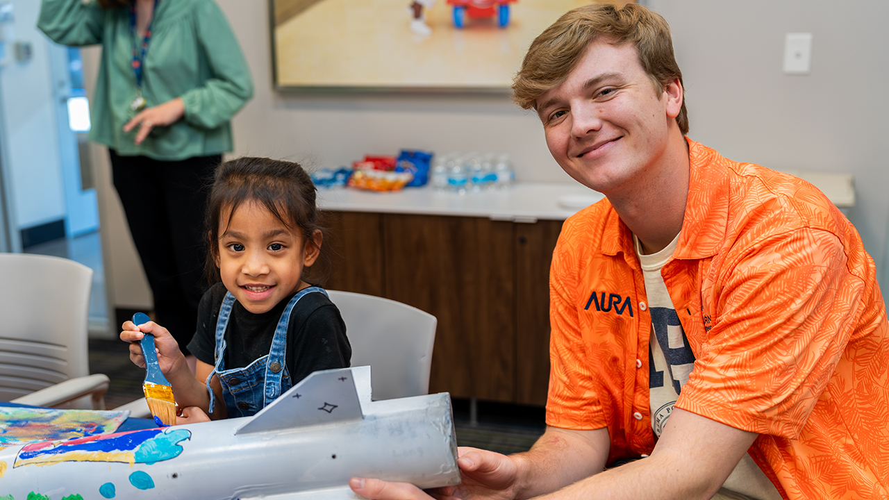 young girl in blue overalls smiles while holding blue paintbrush next to college student in orange shirt holding the end of a rocket.