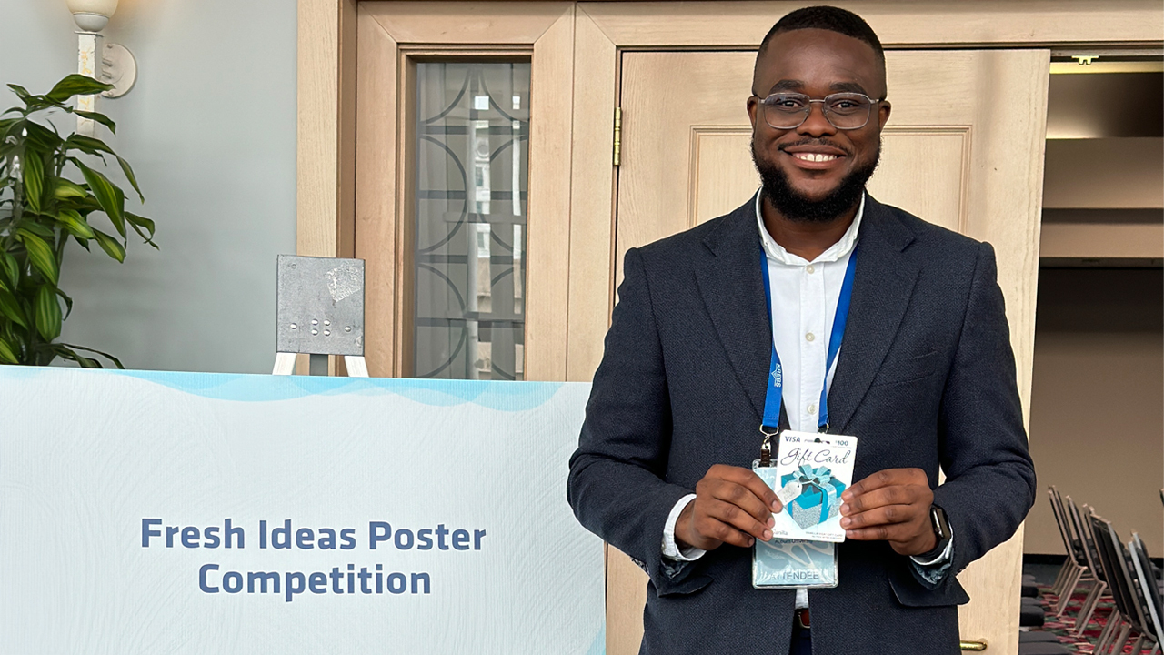 A student in a suit poses for a photo next to a sign that says fresh ideas poster competition.