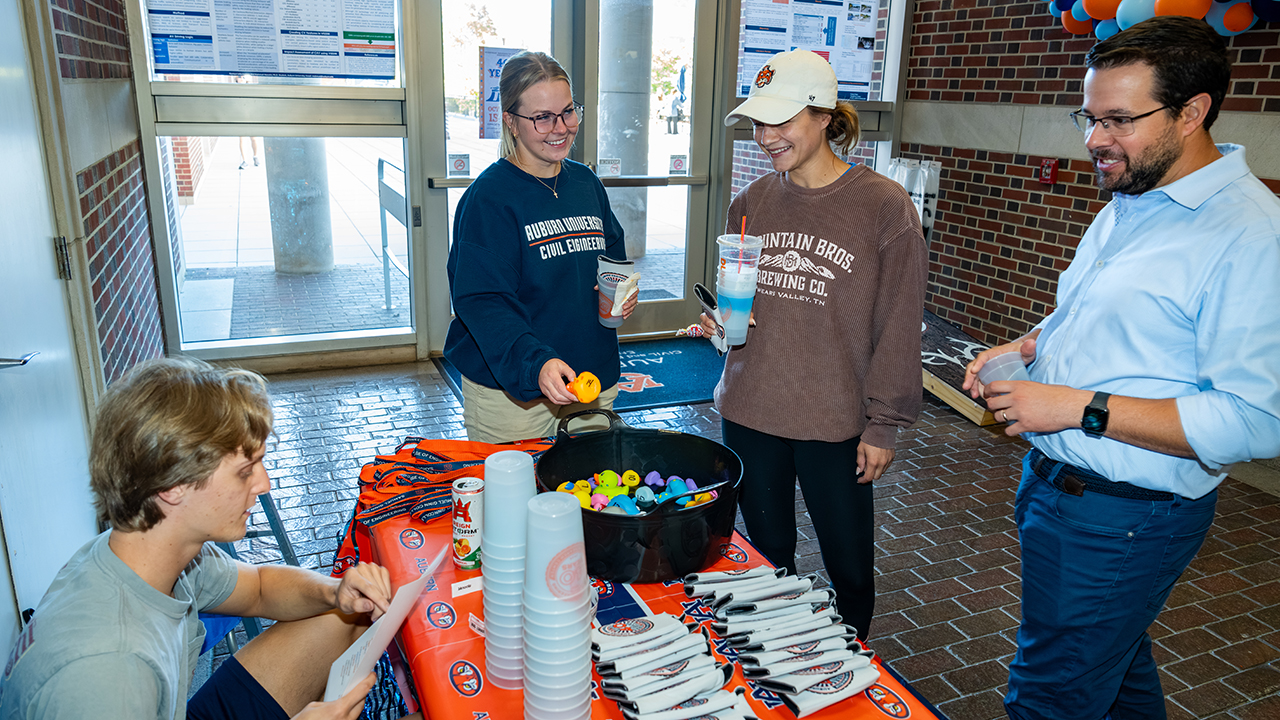 Students gather at an Auburn Engineering outreach table inside a campus building, where one student runs a booth with giveaways like cups and small rubber ducks. Two students holding drinks stop to participate while a faculty member stands nearby, smiling. 