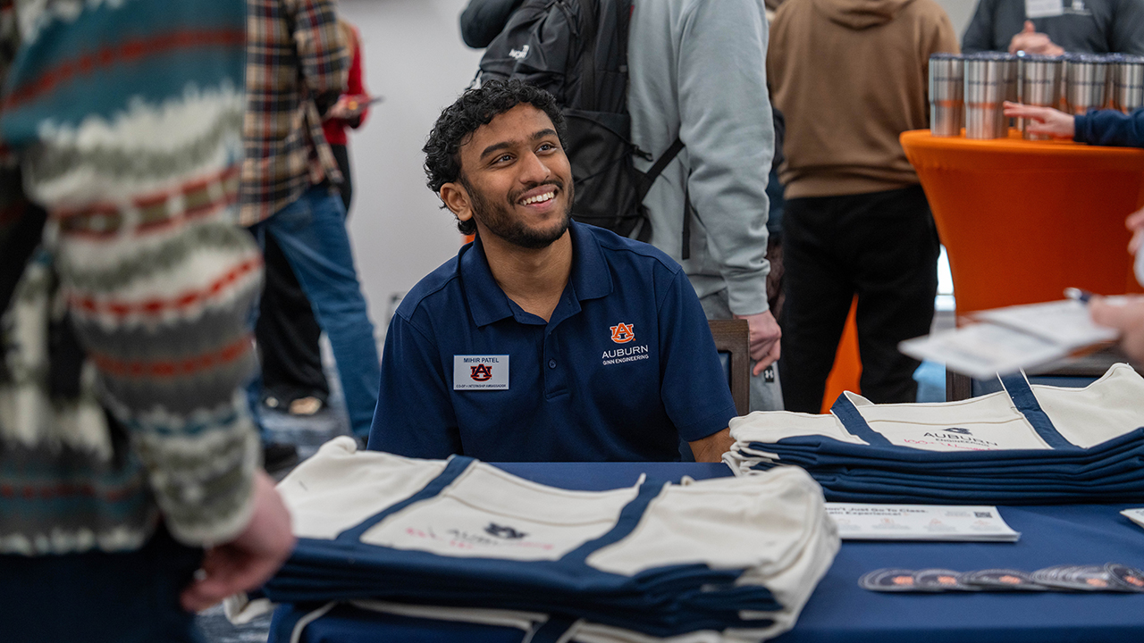 An Auburn Engineering student wearing a navy Auburn polo and name tag smiles while speaking with attendees at a table displaying Auburn tote bags during the Passport to Your Engineering Experience event.