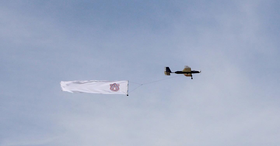 A remote-controlled aircraft flies while towing a banner during the AIAA Design Build Fly competition.