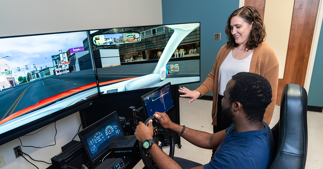 Missie Smith stands beside a participant using a driving simulator with multiple display screens as part of augmented reality and human factors research.