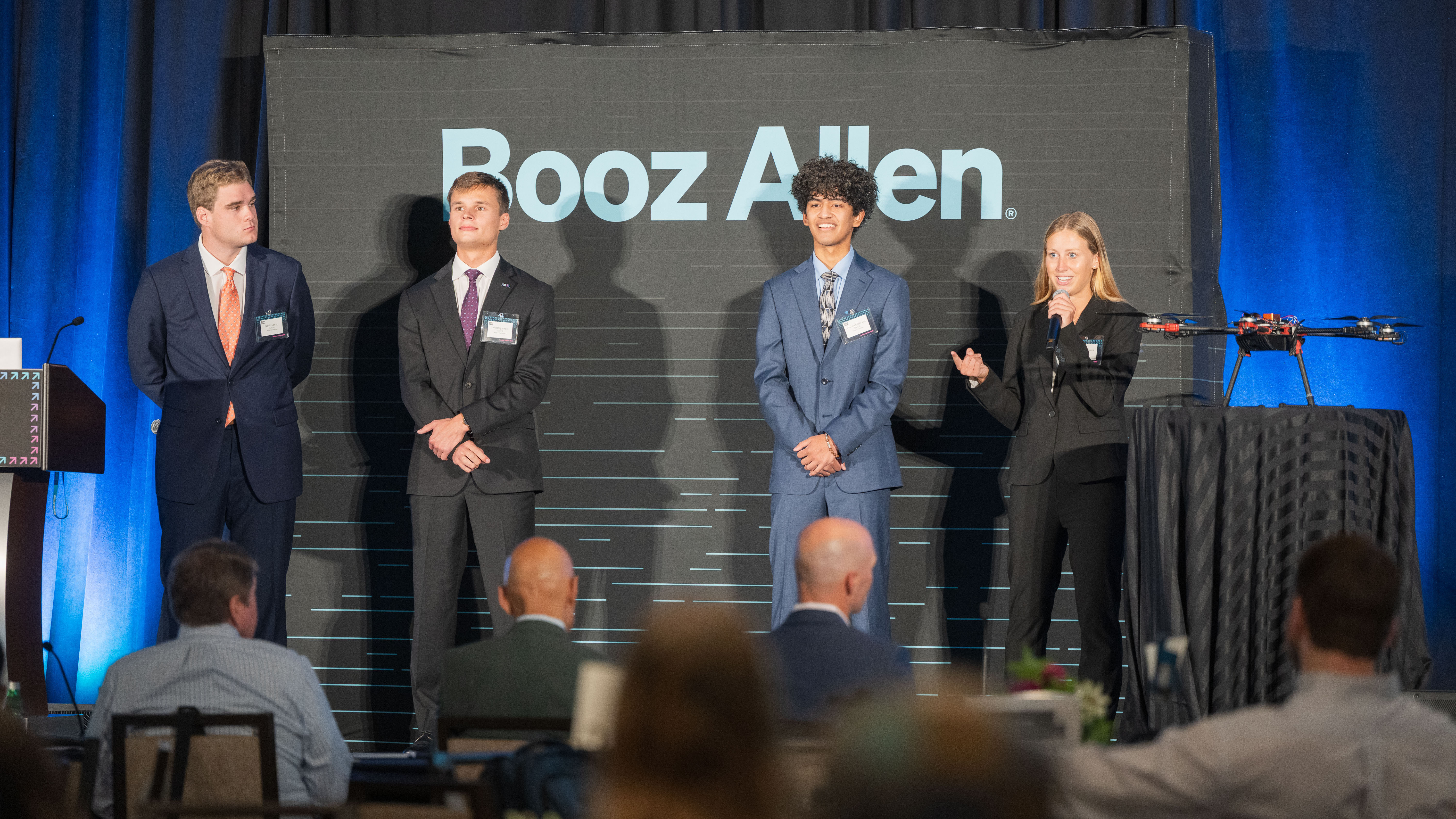  Four students in business attire stand on stage presenting their project at the Booz Allen Summer Games, with Anna Kate Boles speaking into a microphone.