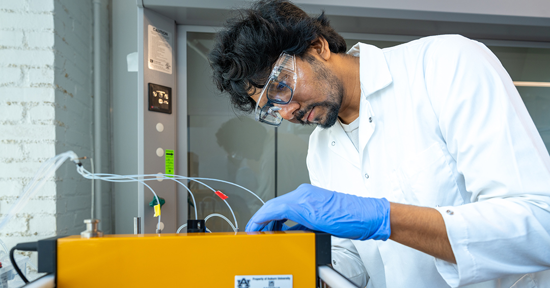 A researcher wearing a white lab coat, safety glasses and blue gloves adjusts laboratory equipment connected to several sample containers.