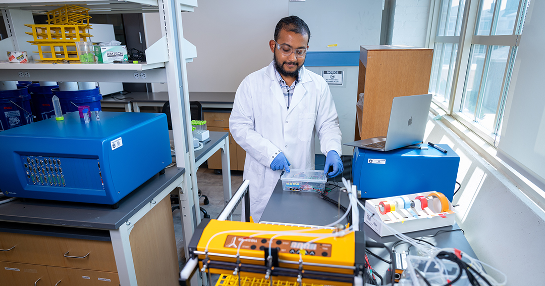 A researcher wearing a white lab coat, safety glasses and blue gloves works with samples and testing equipment at a laboratory bench.