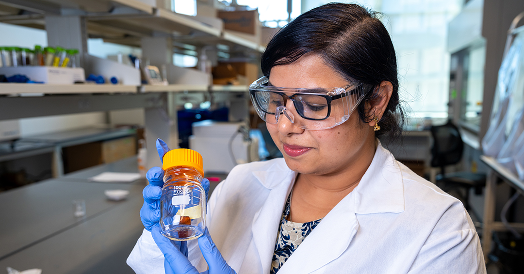 Neha Sharma, wearing a lab coat, safety glasses and blue gloves, holds a glass bottle containing an amber-colored sample in a laboratory.