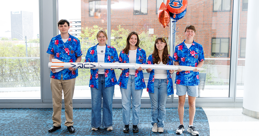 Five Auburn University Rocketry Association students stand indoors holding a model rocket during a visit to Children’s of Alabama, wearing matching blue patterned shirts.